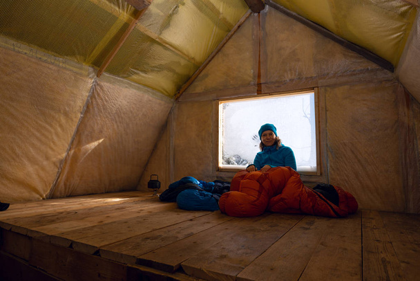 Glückliche Reisende, Frau ruht in der alten Berghütte, sitzt in einem Schlafsack neben einem Fenster, das mit Raureif bedeckt ist, lächelt und blickt in die Kamera. Tolles Abenteuer in der winterlichen Wildnis.  - Foto, Bild