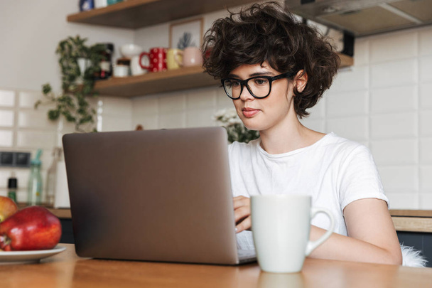 Image of a happy young woman sitting indoors at kitchen using laptop computer. - Foto, Bild