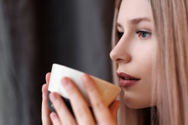 Close-up blonde girl with a cup of hot drink, tea or coffee, at home in a cozy room. - Fotografie, Obrázek
