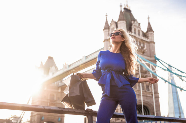 The happy beautiful young blonde woman in sunglasses standing against the background of the bridge and holding shopping bags in hand - Foto, afbeelding