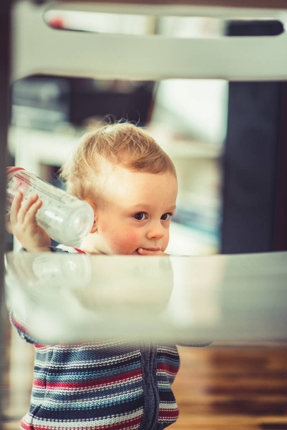 Um bebê doce está de pé e segurando sua garrafa vazia
 - Foto, Imagem