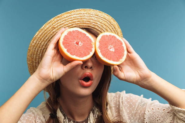 Image of a happy young excited beautiful woman isolated over blue wall holding citrus wearing hat covering eyes. - Фото, зображення