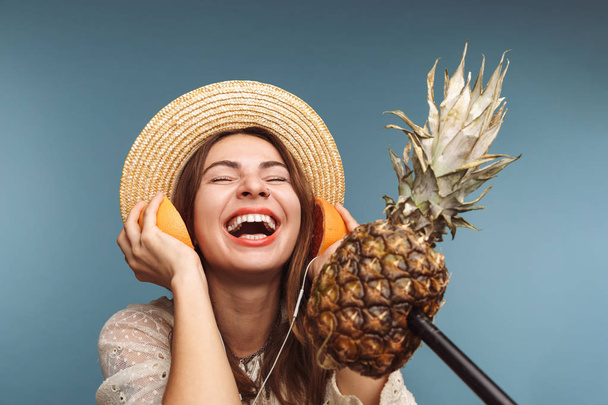 Image of excited young beautiful woman isolated over blue wall holding citruses near ears as a headphones and singing to pineapple as a mic. - Fotoğraf, Görsel