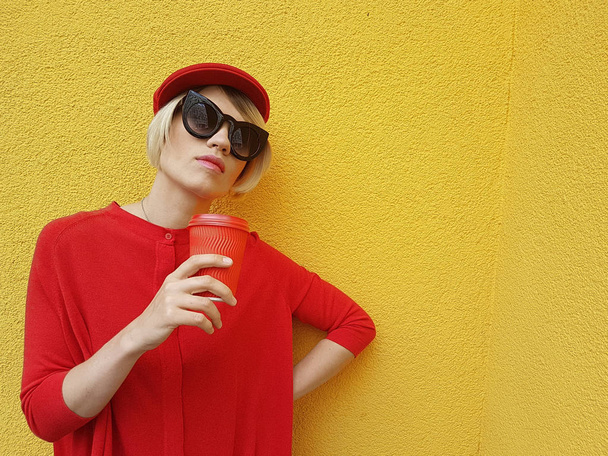 Female model in long red sweater and red hat with red paper cup standing on yellow background. Outdoor photo of beautiful caucasian woman in sweater drinks coffee in front of cafe - Foto, Imagem
