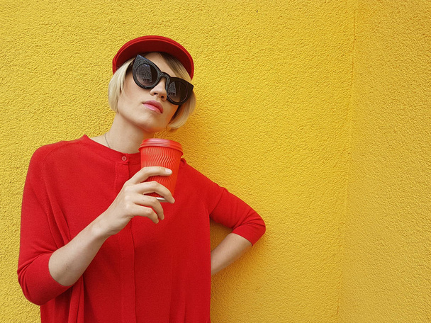 Vrouwelijke model in lange rode trui en rode hoed met rode papier beker staande op gele achtergrond. Buiten foto van mooie Kaukasische vrouw in trui drinkt koffie tegenover café - Foto, afbeelding