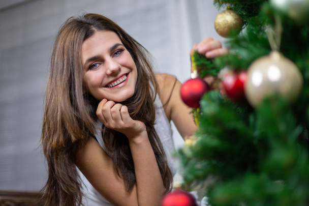Beautiful young female smiling and looking at camera while hanging small bauble on Christmas tree - Valokuva, kuva