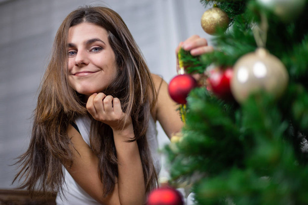 Beautiful young female smiling and looking at camera while hanging small bauble on Christmas tree - Zdjęcie, obraz