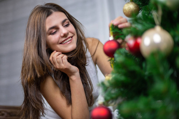 Beautiful young female smiling and hanging small bauble on Christmas tree - Fotoğraf, Görsel