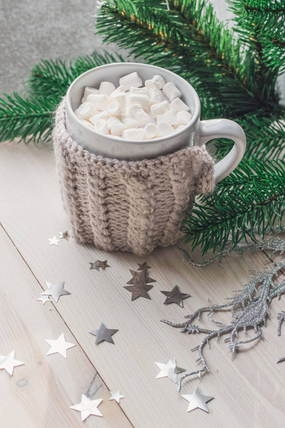 Xícara branca com marshmallows em uma jaqueta de malha quente em um fundo de madeira leve com uma árvore de Natal e estrelas argênteas. espaço para texto
 - Foto, Imagem