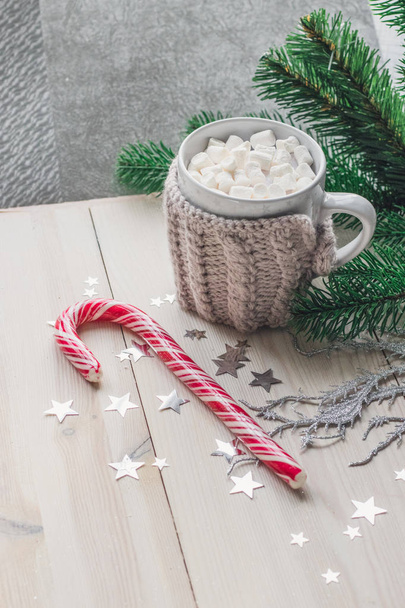 weiße Tasse mit Marshmallows in warmer Strickjacke auf hellem Holzgrund mit Weihnachtsbaum und silbernen Sternen. Raum für Text - Foto, Bild