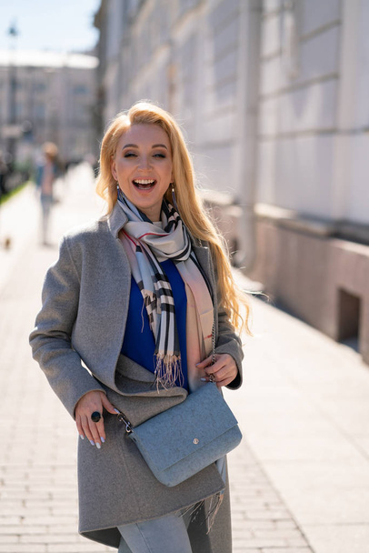 charming blonde girl with long hair in a trendy gray coat with beige checkered scarf smiles and walks around the city like a tourist and enjoys a Sunny spring day - Foto, Imagen