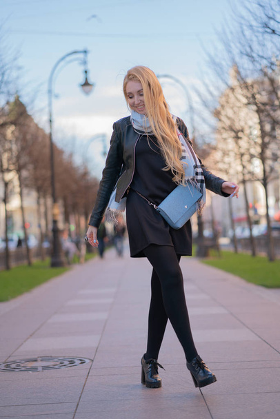 lovely blonde woman in a leather jacket, black dress and with a checkered beige scarf walks around the City and enjoys life on the positive - Fotoğraf, Görsel