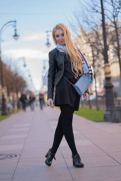 Lovely білявка в шкіряна куртка, чорні сукні і з шашечками бежевий шарф прогулянки по місту і насолоджується життям на позитивні - Фото, зображення