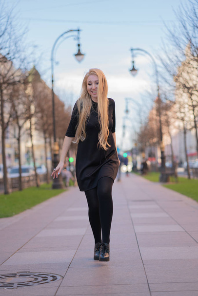 lovely blonde woman in a leather jacket, black dress and with a checkered beige scarf walks around the City and enjoys life on the positive - Zdjęcie, obraz