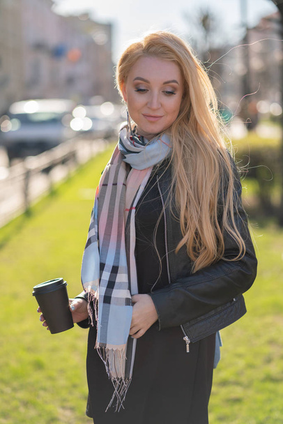 cute blonde woman in a jacket with a checkered beige scarf walks around the green city with a Cup of coffee in her hands - Foto, immagini