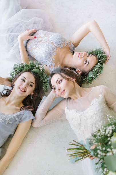 Pretty bride with bridesmaids are lying on the white floor. Bridedmaids in the same grey color rustic dress and wreath of pine and eucalyptus on their heads. Wedding morning. - Photo, image