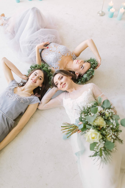 Top view of bride with female friends lying on the floor and smiling. Three young women lying on bed at home. Wedding morning before ceremony - Photo, image
