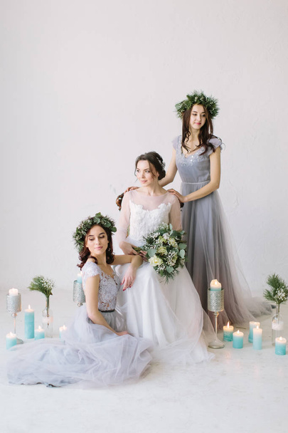 Beautiful bride in a white wedding dress holding bouquet and sitting on the vintage chair with bridesmaids in blue grey dresses and wreathes. Wedding decor and candles on the background - Photo, image
