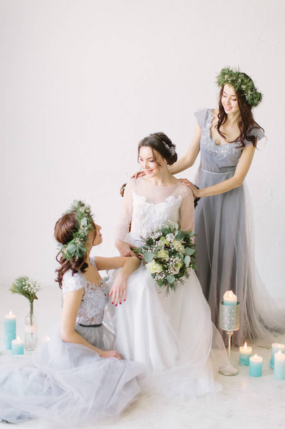 Beautiful bride in a white wedding dress holding bouquet and sitting on the vintage chair with bridesmaids in blue grey dresses and wreathes. Wedding decor and candles on the background - Photo, Image
