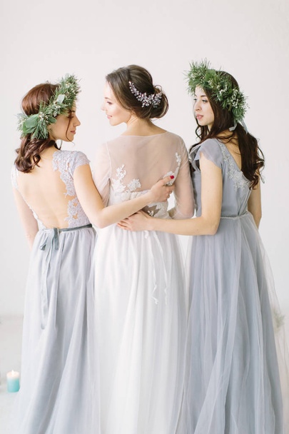 Wedding. Pretty bride in a white dress standing and embracing bridesmaids in gray blue dresses and wreathes - Фото, изображение