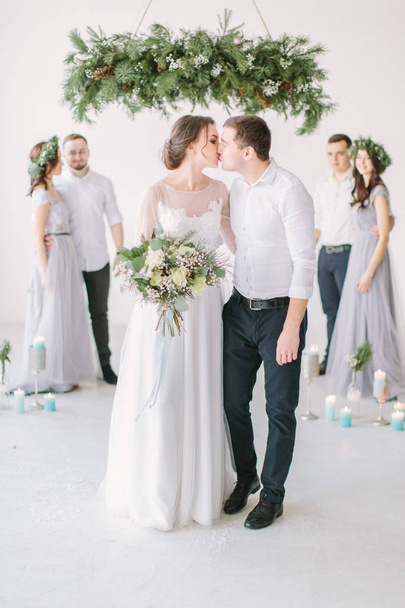 Handsome brunette groom kissing beautiful bride in wedding dress with bouquet and groommen and bridesmaids in background - Zdjęcie, obraz