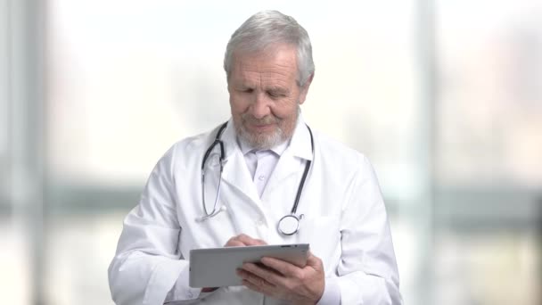 Médico anciano sonriente con comprimido de pc. Alegre médico caucásico utilizando tableta de ordenador sobre fondo borroso abstracto
. - Imágenes, Vídeo