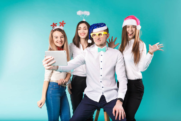Новогодняя тема зимнего офиса сотрудников компании. Группа 4 молодых людей Кавказа бизнес улыбка праздник смешные шляпы аксессуары очки сфотографировать себя селфи планшет синий фон
 - Фото, изображение