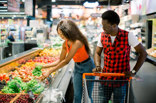 smiling african american couple with shopping trolley choosing vegetables in supermarket - Foto, Imagen