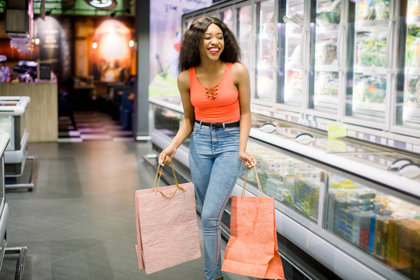 Αρκετά χαμογελαστός αφρικανικό αμερικανικό κορίτσι σε casual ρούχα κρατώντας ροζ χάρτινες σακούλες στα σούπερ μάρκετ. Εμπορική ιδέα και έννοια - Φωτογραφία, εικόνα
