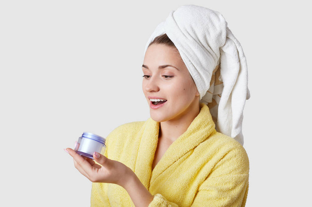 Joyful young European woman holds bottle with cream, looks happily at new cosmetic product, has white towel on head, stands against white studio background, has beauty treatments in bathroom - Photo, image