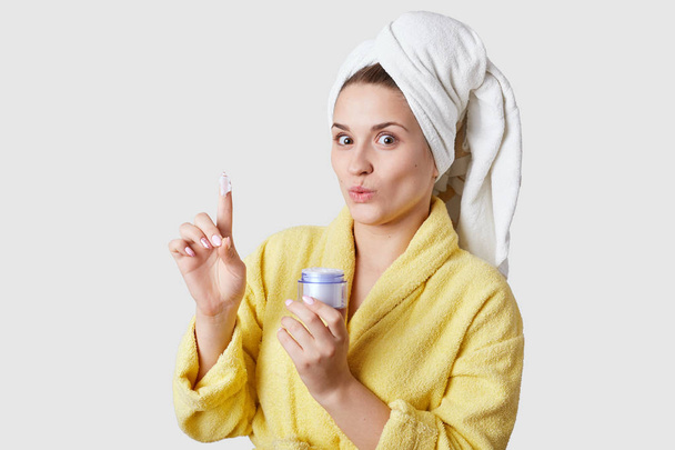 Skin care concept. Beautiful young lady holds cosmetic cream, has joyful surprised facial expression, wears yellow bathrobe, isolated over white studio wall. Face treatment. Moisture for skin - Valokuva, kuva