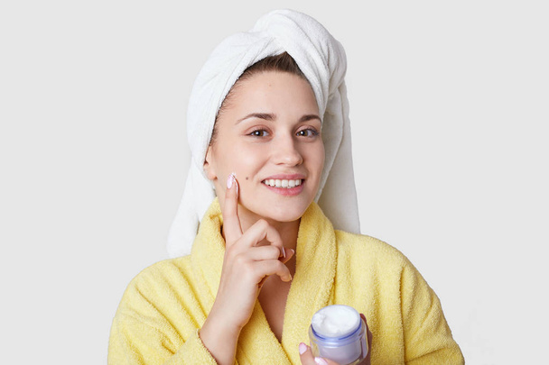 Smiling woman keeps fore finger on face, uses cosmetic cream for staying young and fresh, wears white towel and bathrobe, models against white background. Body care at home. Mask for skin face - Φωτογραφία, εικόνα