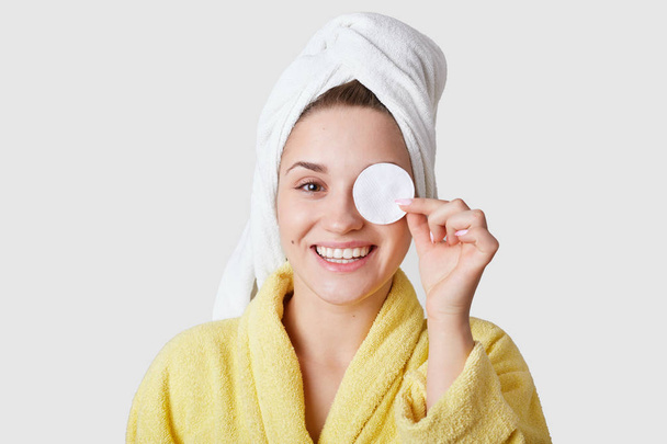 Studio shot of pleasant looking young smiling European woman covers eye with cotton disk, has white towel on head, takes care of skin, models against white studio wall. Beauty procedure at home - Foto, Imagem