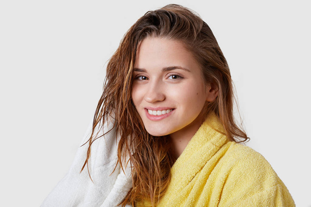 Clean smiling young female looks happily at camera, has tender smile, wet dark hair, wipes with towel, dressed in comfortable bathrobe, stands against white background. Human feelings, beauty concept - Foto, Bild