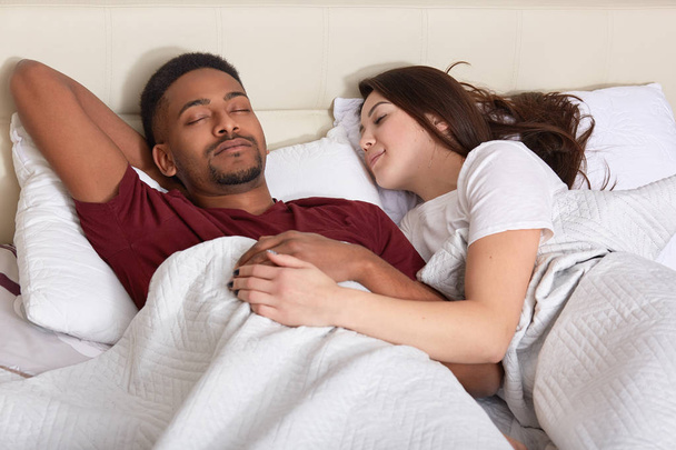 People, bed time concept. Interracial affectionate couple have healthy sleep in bed, enjoy relaxation, enjoy good rest, pose in bedroom. African American young man takes nap with Caucasian wife. - Valokuva, kuva