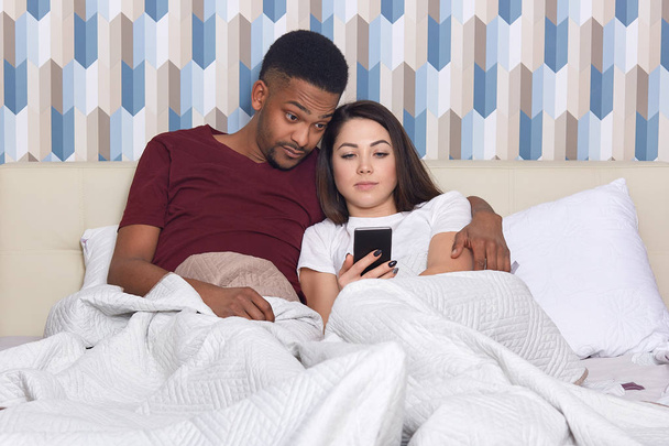 Surprised dark skinned man embraces his wife, pose together in comfortable bed, view photos or read news in internet on cell phone, enjoy comfort, awake in morning. Bed time, interracial relationship - Φωτογραφία, εικόνα