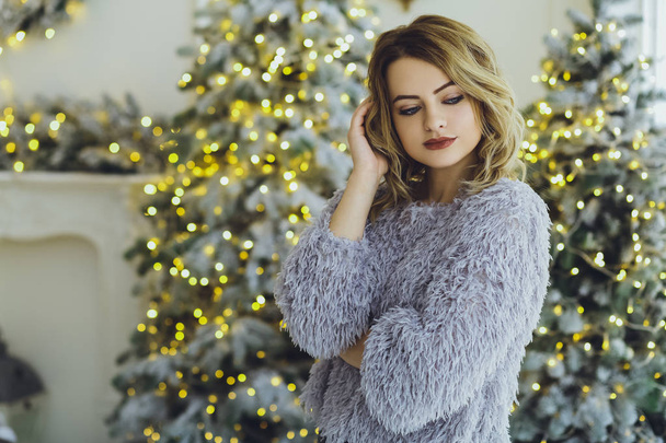 Young beautiful blonde woman in a gray sweater with a gift on hands near the New Year, Christmas tree with gifts - Фото, изображение