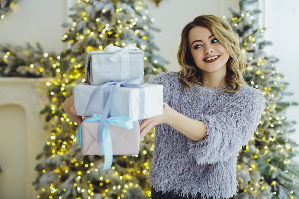Young beautiful blonde woman in a gray sweater with a gift on hands near the New Year, Christmas tree with gifts - Fotó, kép
