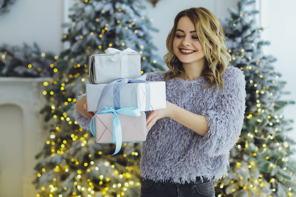 Young beautiful blonde woman in a gray sweater with a gift on hands near the New Year, Christmas tree with gifts - Photo, image