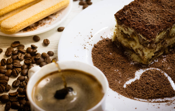 Tiramisu Italiaans dessert met hart van geraspte chocolade op wit bord met kopje koffie splash druppels, savoiardi koekjes en verspreide koffiebonen - Foto, afbeelding