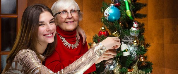 new year. senior grandma and young caucasian woman, teenager near christmas tree. granddaughter, teen hugging, embraces old grandmother. happy company. cheerful girl smiles, laugh. festive spirit - Foto, Bild