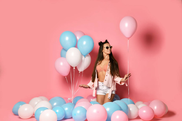 Portrait of pretty joyful young woman holding box with gift present on pastel pink background with colorful air balloons. - 写真・画像