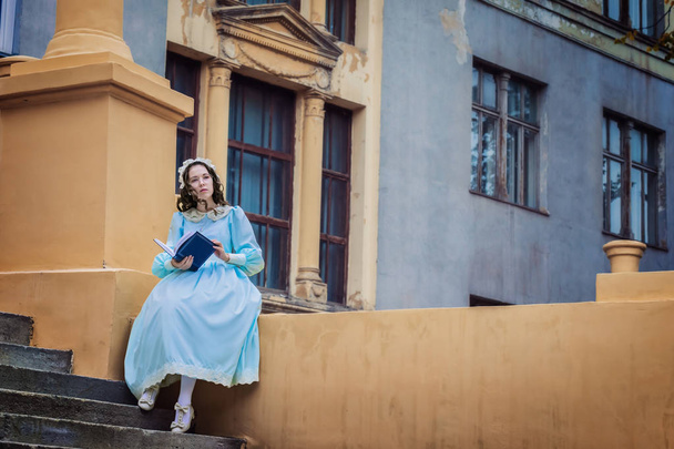 Young, doordachte meisje in een blauwe vintage jurk leest een liefdesverhaal op de trappen van het kasteel - Foto, afbeelding