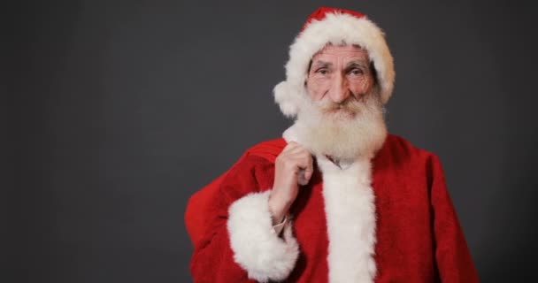 Old, kind santa with long white beard showing red bag full of chistmas gifts, isolated shoot in gray background - Video