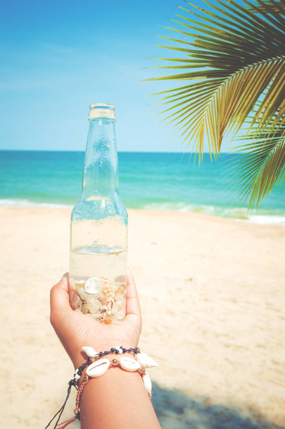 Relaxation and Leisure in summer - Young tanned woman hand holding bottle with seashell at tropical beach in summer. vintage color tone effect - Foto, Bild