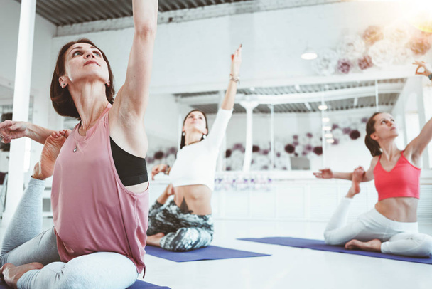 Close-up view on strong healthy woman practicing yoga exerciese on fitness mat in white sport class. Adult fit female doing stretching early morning indoor on background people - Foto, Imagen