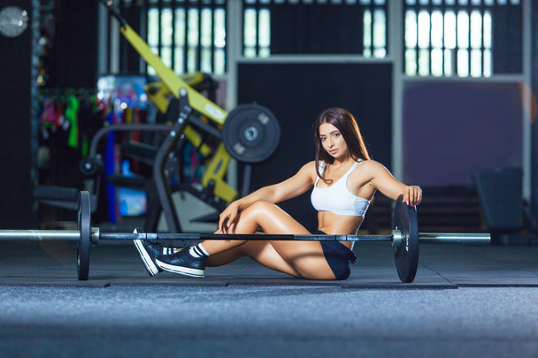 Fit long-legged woman sitting on the floor at gym near barbell - Фото, зображення