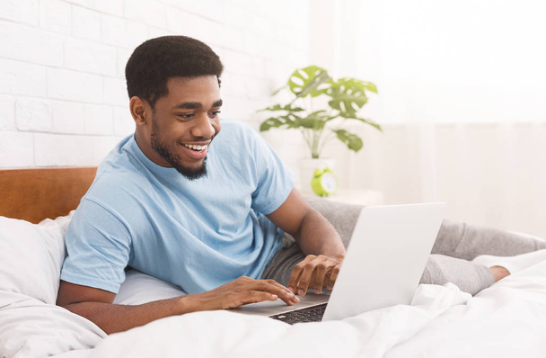 Zwarte man in bed liggen en kijken naar iets op laptop - Foto, afbeelding