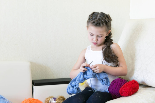 The girl knits with knitting and thread. A little girl learns to knit clothes sitting on the couch. - Foto, afbeelding