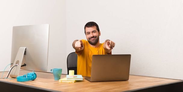 Mężczyzna pracujący z laptot w biurze wskazuje palcem na Ciebie jednocześnie uśmiechając się - Zdjęcie, obraz